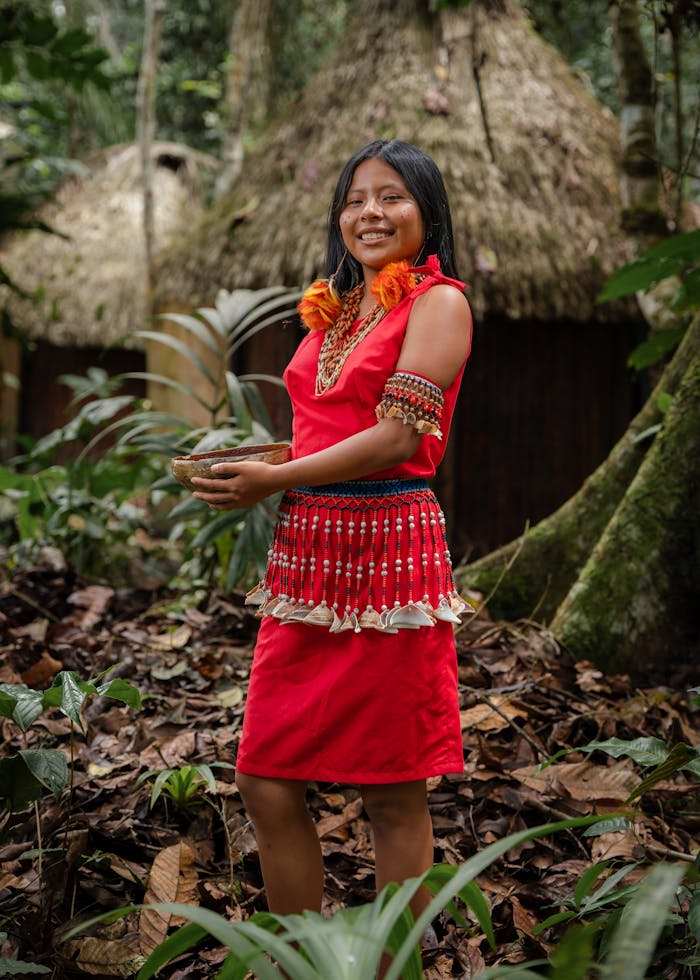 A smiling indigenous woman wearing traditional red attire in a lush forest in San Martín, Peru.